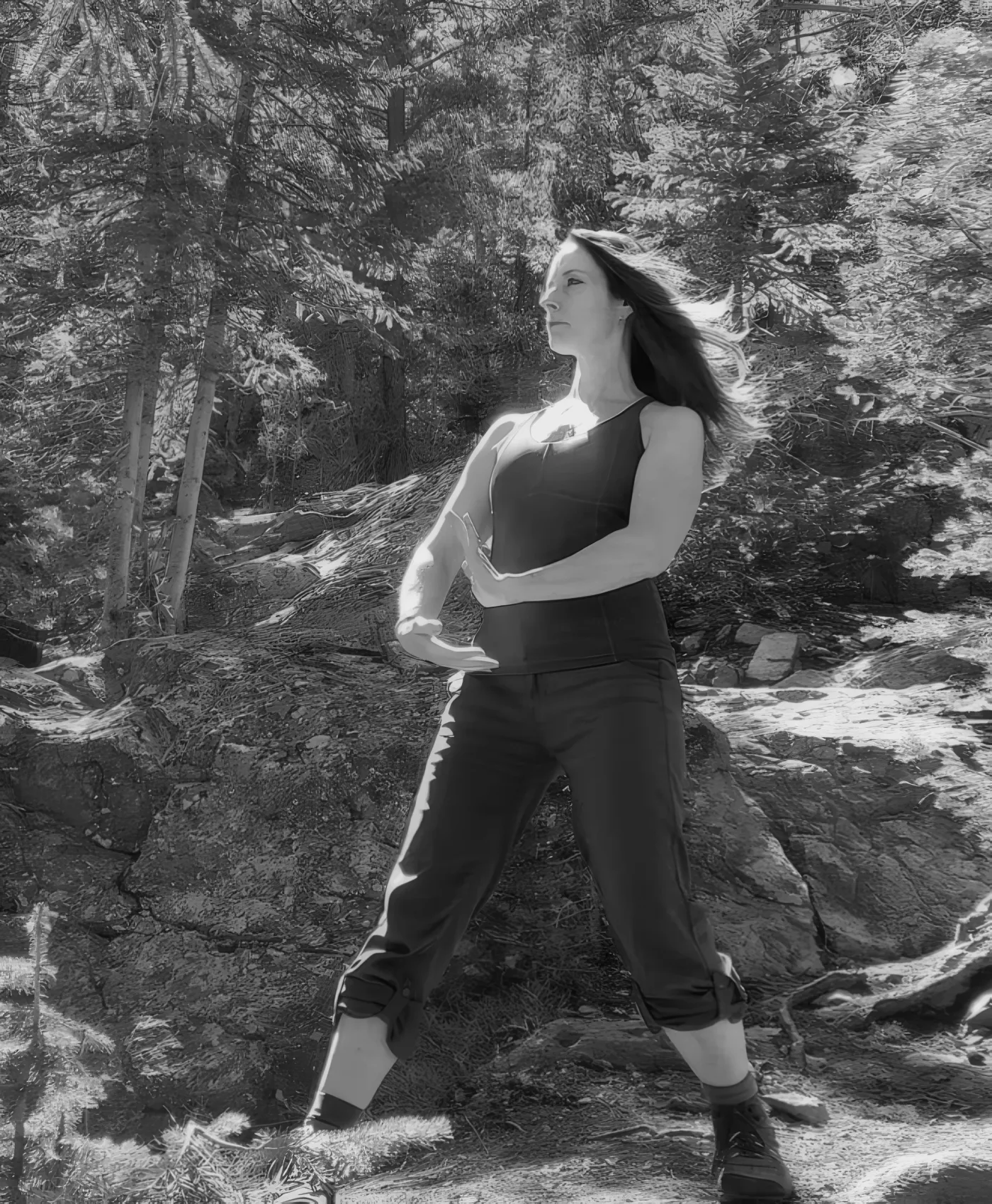 Isshaela in a Qigong stance among pines, in black and white, her hair caught by the wind